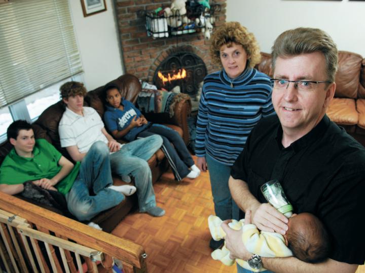 Living room with many children and two adults, man is bottle-feeding a baby