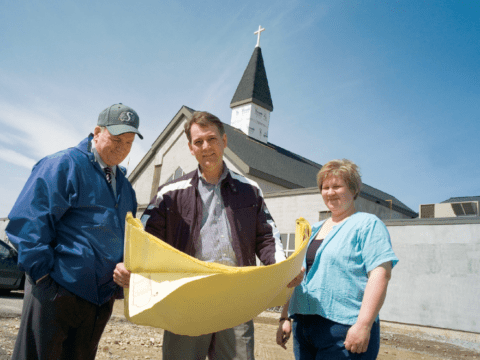Two white men and a white woman looking at blueprint plans in front of partly constructed steepled church