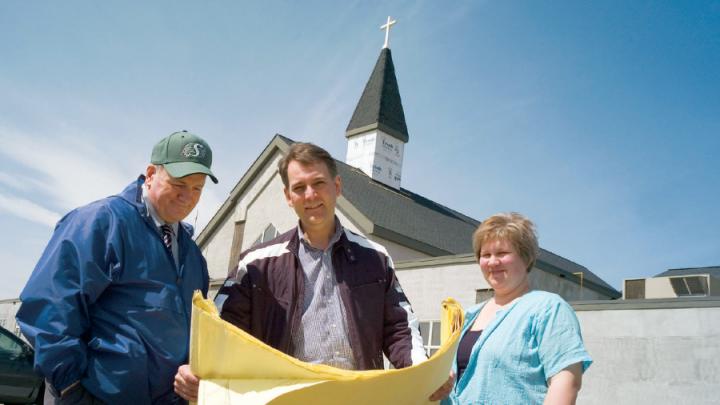 Two white men and a white woman looking at blueprint plans in front of partly constructed steepled church
