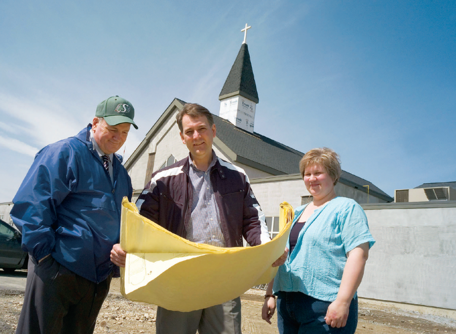 Two white men and a white woman looking at blueprint plans in front of partly constructed steepled church