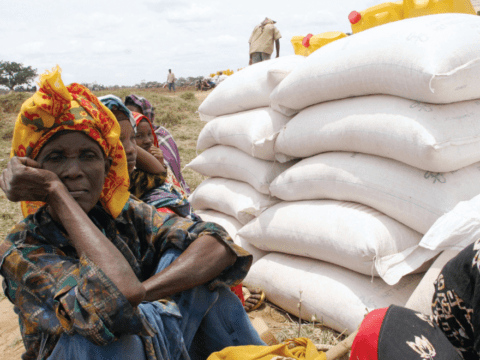 Black women in headscarves sit on ground next to bags of grain