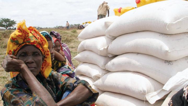 Black women in headscarves sit on ground next to bags of grain
