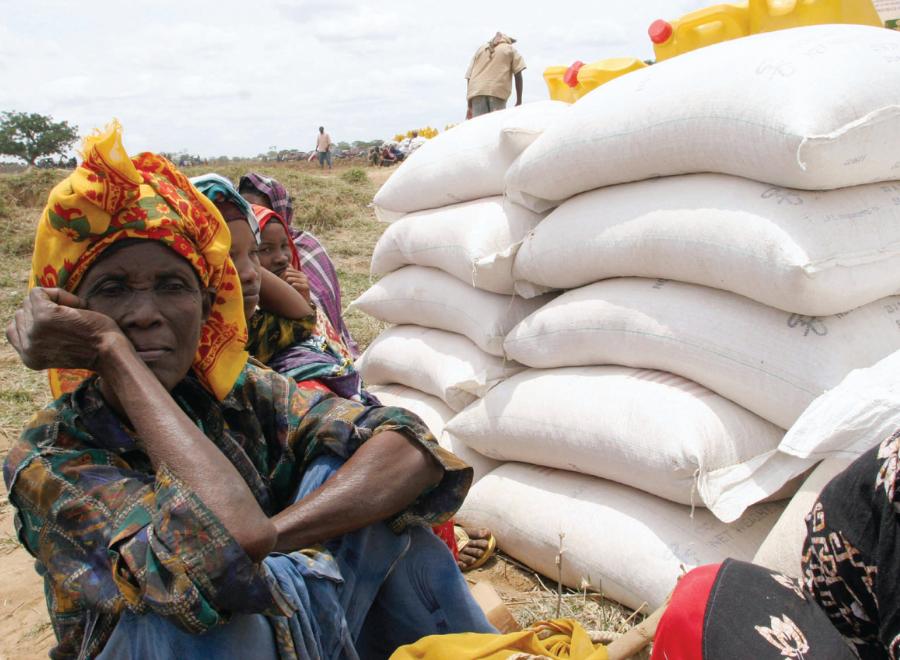 Black women in headscarves sit on ground next to bags of grain