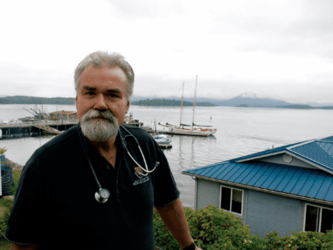 White-bearded man with stethoscope around his neck standing in front of boat harbour
