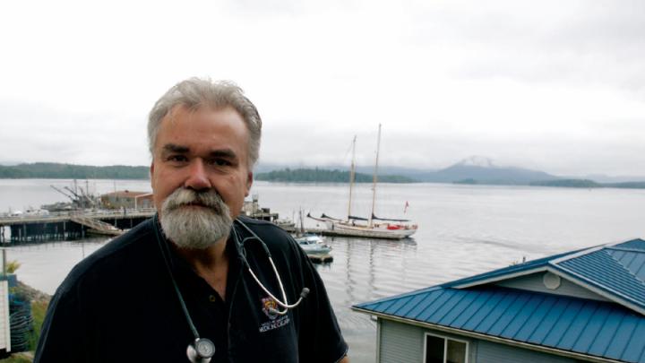 White-bearded man with stethoscope around his neck standing in front of boat harbour