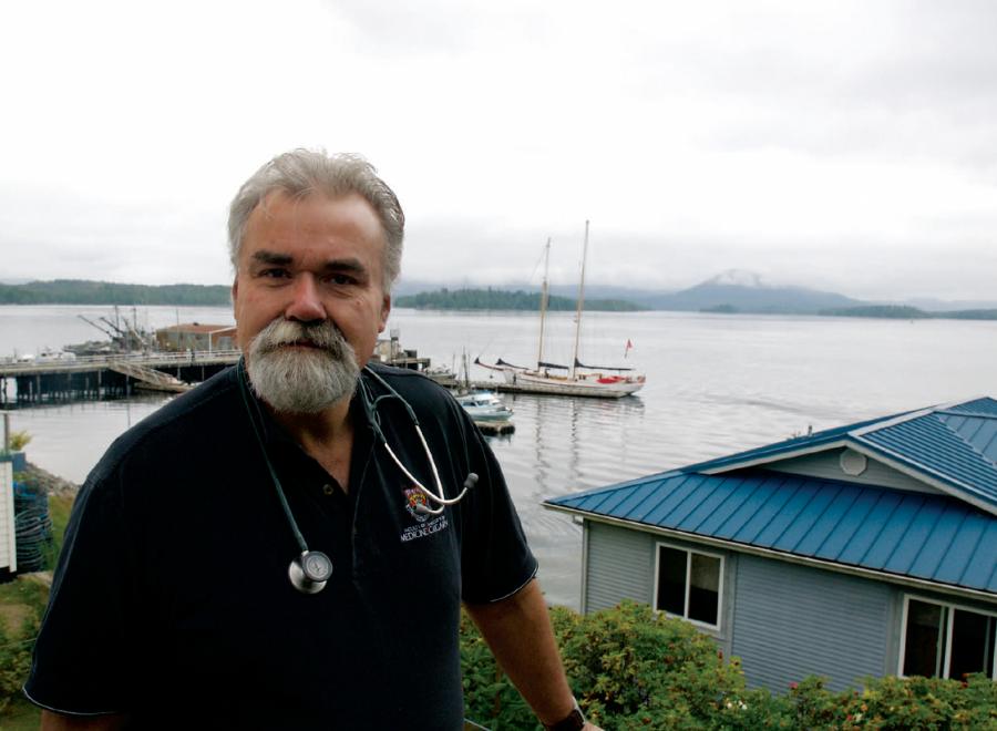 White-bearded man with stethoscope around his neck standing in front of boat harbour