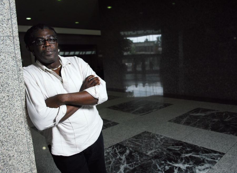 Black man in white shirt with arms crossed leaning against wall