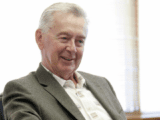 White-haired white man in suit jacket and open-necked white shirt