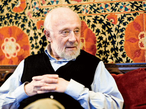 White-haired white man sitting in front of colourful backdrop