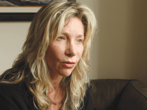 White woman with blonde medium layered cut hair and hazel eyes staring into the distance. She has no smile.