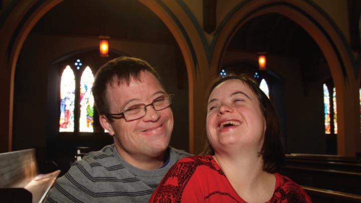Disabled couple in church smiling with each other in the dimly lit pew. Stained glass windows are in the background