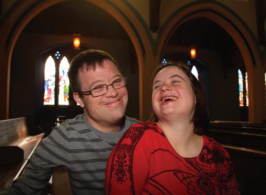 Disabled couple in church smiling with each other in the dimly lit pew. Stained glass windows are in the background