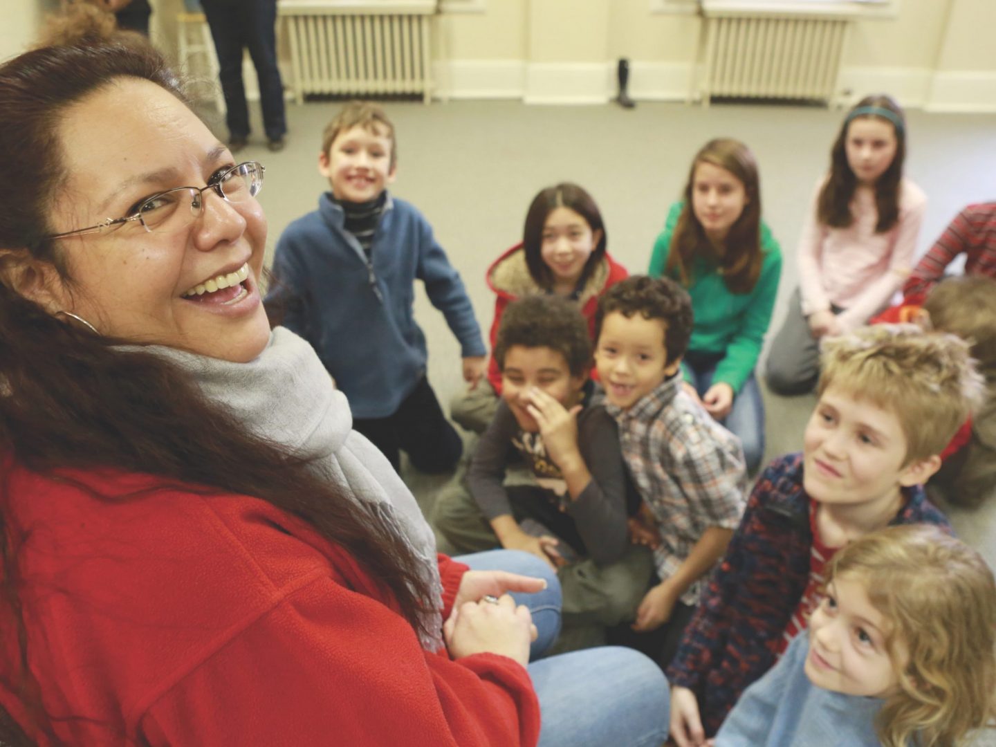 Woman in red jacket looks at camera, circle of children on floor smiling