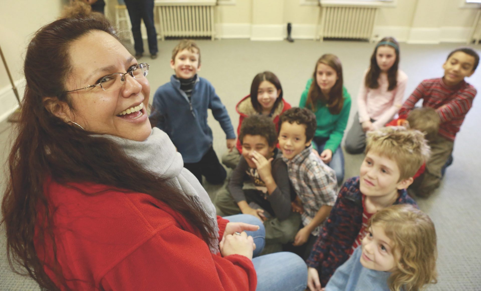 Woman in red jacket looks at camera, circle of children on floor smiling