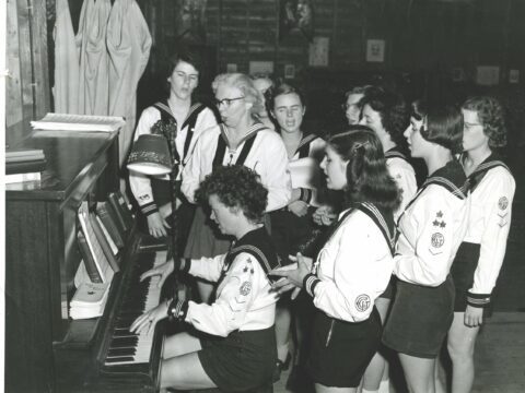 Archival photo of young women singing around piano