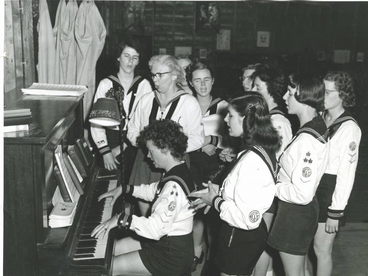 Archival photo of young women singing around piano