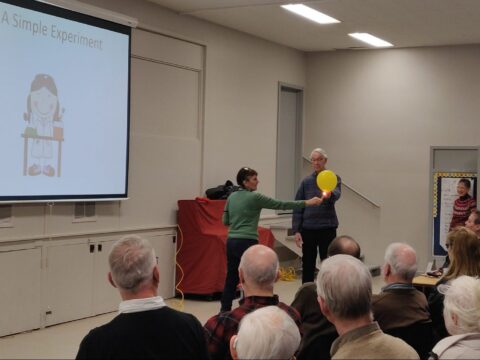 A woman holds a flame against a balloon held by a man. They are in a large room, watched by about 20 people sitting in chairs.