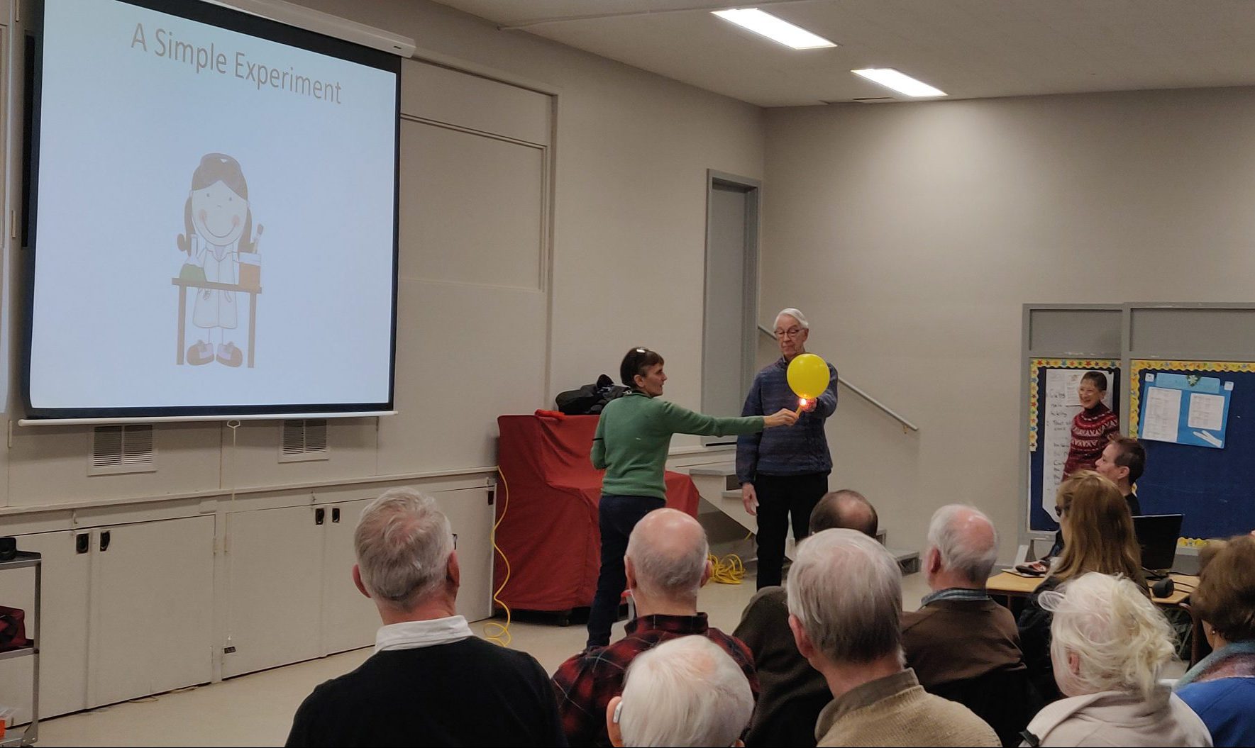 A woman holds a flame against a balloon held by a man. They are in a large room, watched by about 20 people sitting in chairs.