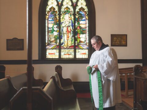 Anglican priest Theo Robinson stands in front of a church pew.