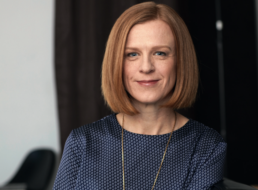 Woman with ginger hair cut into a bob. She is wearing a blue blouse with a long gold chain.