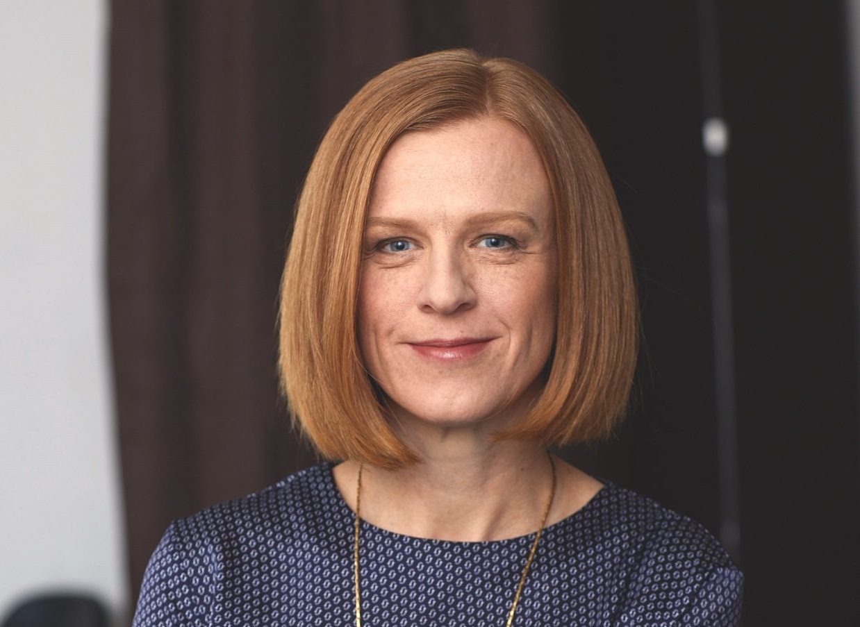Woman with ginger hair cut into a bob. She is wearing a blue blouse with a long gold chain.