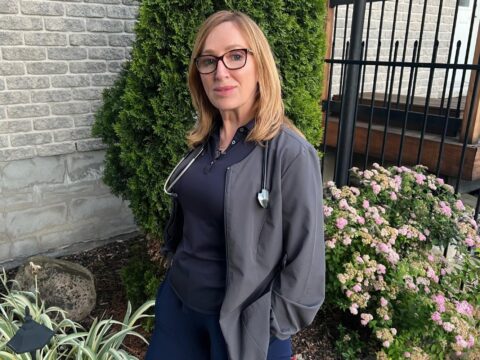 Debra Lefebvre stands in dark blue scrubs with a stethoscope hanging around her neck, in front of a small garden bed