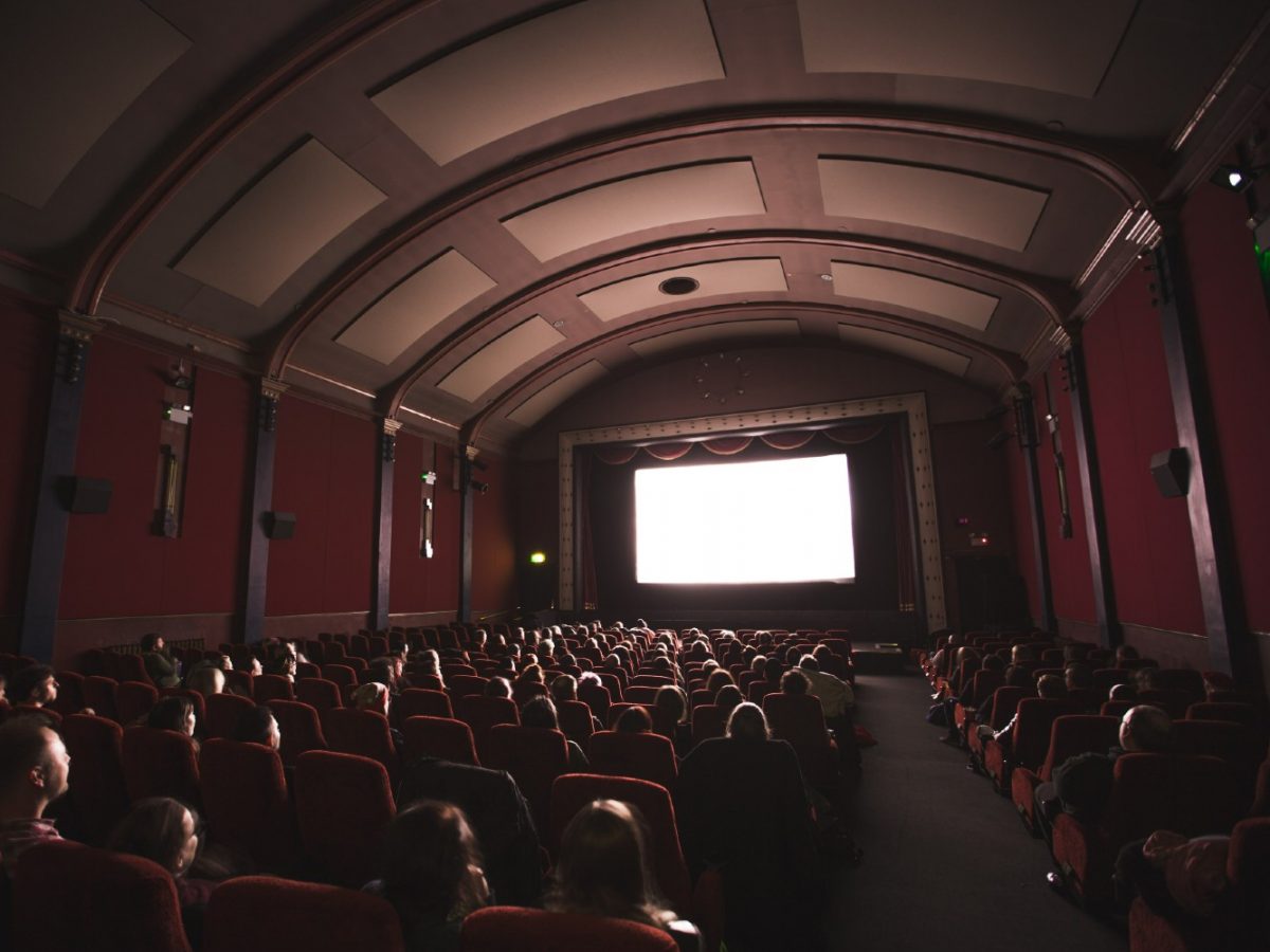 People sit in the dark in a maroon movie theatre, facing a bright white screen