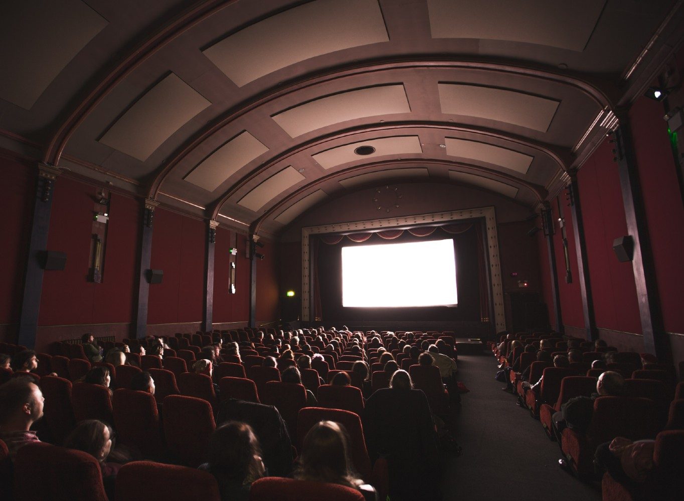 People sit in the dark in a maroon movie theatre, facing a bright white screen