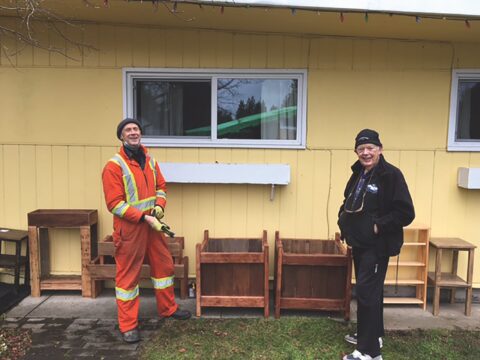 two men stand in front of small building with some small wooden shelves between them