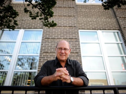 White man with clasped hands in front of building