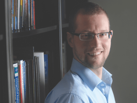 young white man with glasses and beard looking at a camera in front of bookshelf