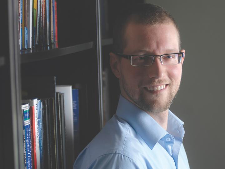young white man with glasses and beard looking at a camera in front of bookshelf