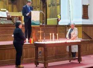 Three people in a church. A man is reading at a podium while two women stand in front of him lighting candles.
