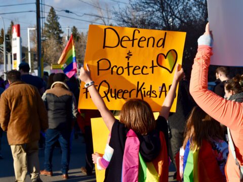 Pro-2SLGBTQ+ protesters march while one holds a pride flag and a yellow sign that reads "Defend & Protect Queer Kids."