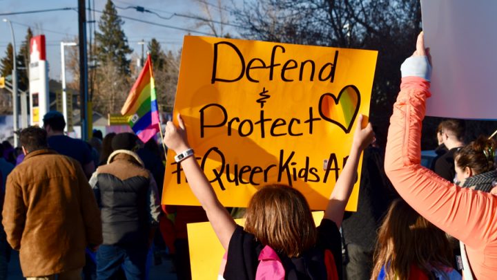 Pro-2SLGBTQ+ protesters march while one holds a pride flag and a yellow sign that reads "Defend & Protect Queer Kids."