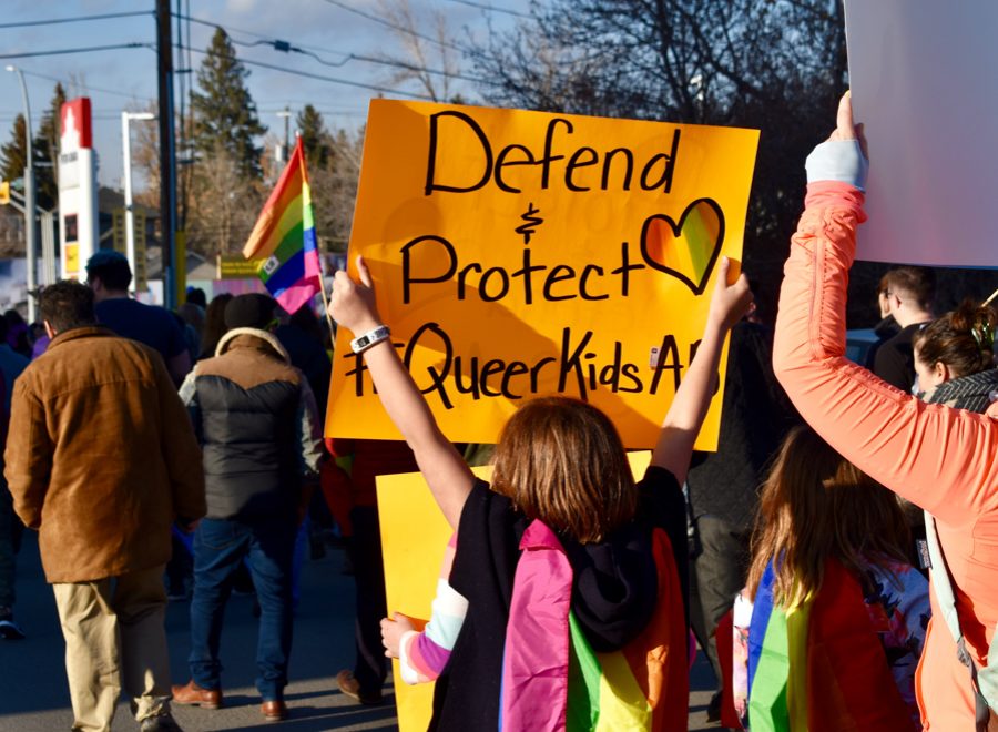 Pro-2SLGBTQ+ protesters march while one holds a pride flag and a yellow sign that reads "Defend & Protect Queer Kids."