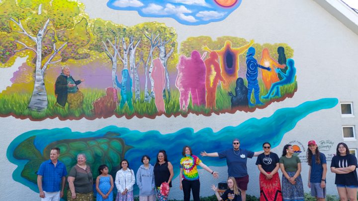 A group of just over a dozen people, mostly youths, are standing in front of a building with a mural on it. The mural features an elder sitting down and speaking to colourful silhouettes of children while surrounded by birch trees and sunny skies. Underneath, Grandmother Turtle is swimming.