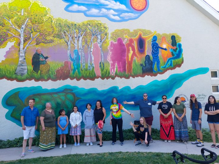 A group of just over a dozen people, mostly youths, are standing in front of a building with a mural on it. The mural features an elder sitting down and speaking to colourful silhouettes of children while surrounded by birch trees and sunny skies. Underneath, Grandmother Turtle is swimming.