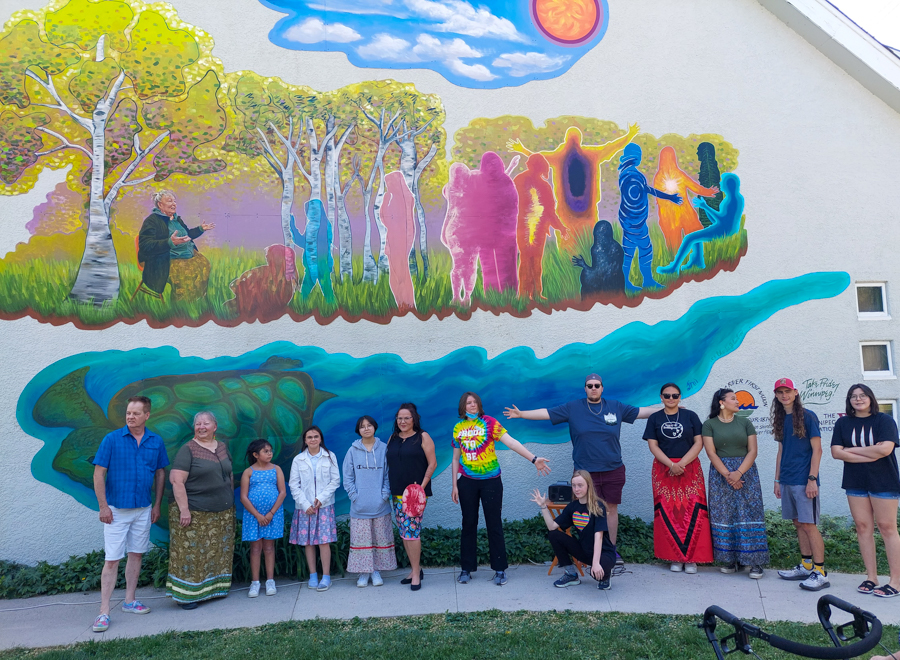 A group of just over a dozen people, mostly youths, are standing in front of a building with a mural on it. The mural features an elder sitting down and speaking to colourful silhouettes of children while surrounded by birch trees and sunny skies. Underneath, Grandmother Turtle is swimming.