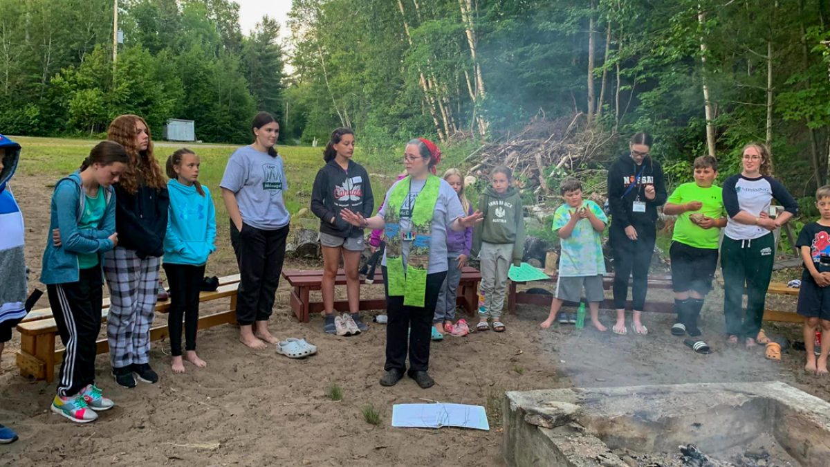 Children and a camp counsellor are seen standing in front of a campfire.