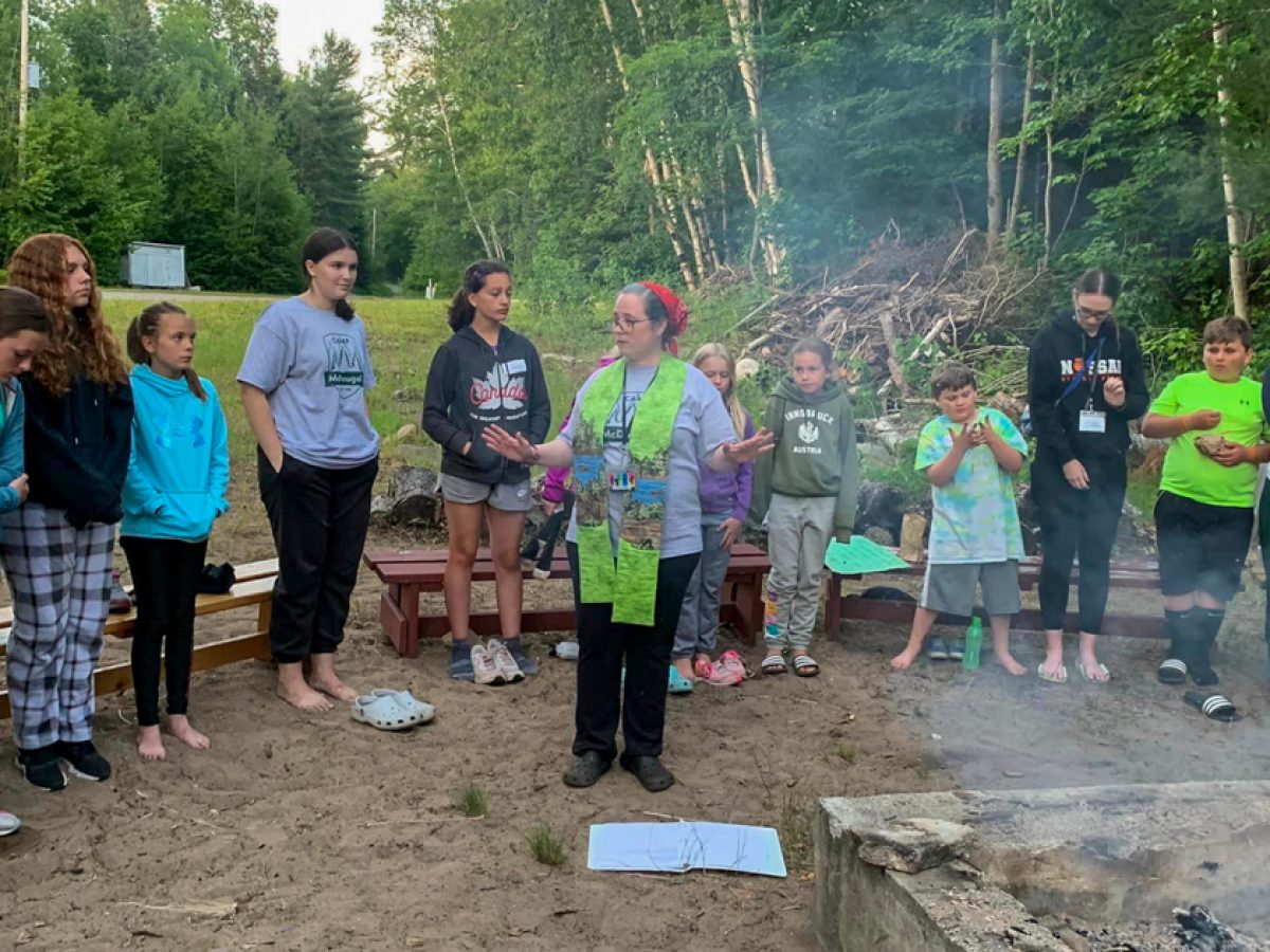 Children and a camp counsellor are seen standing in front of a campfire.
