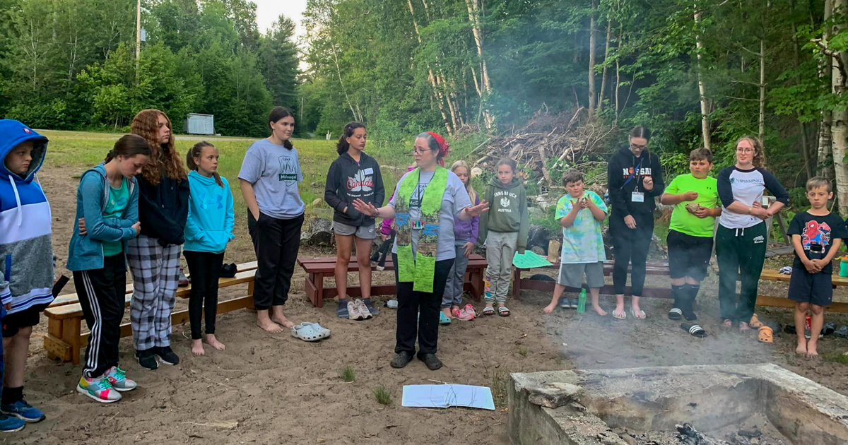 Children and a camp counsellor are seen standing in front of a campfire.