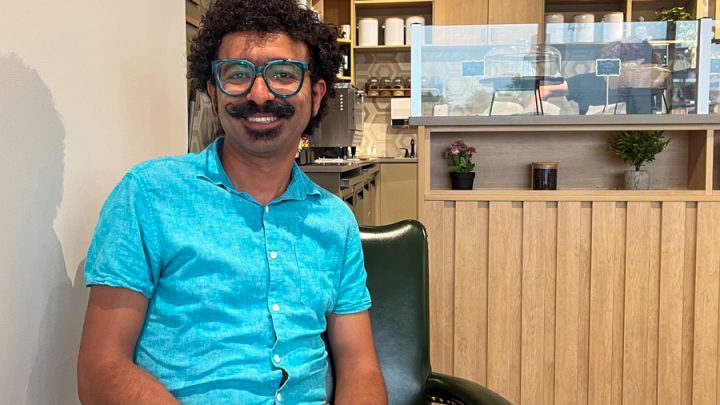 A brown-skinned man with glasses, black curly hair and a moustache is sitting on a chair in a coffee shop smiling. He is wearing a teal shirt and light red shorts.