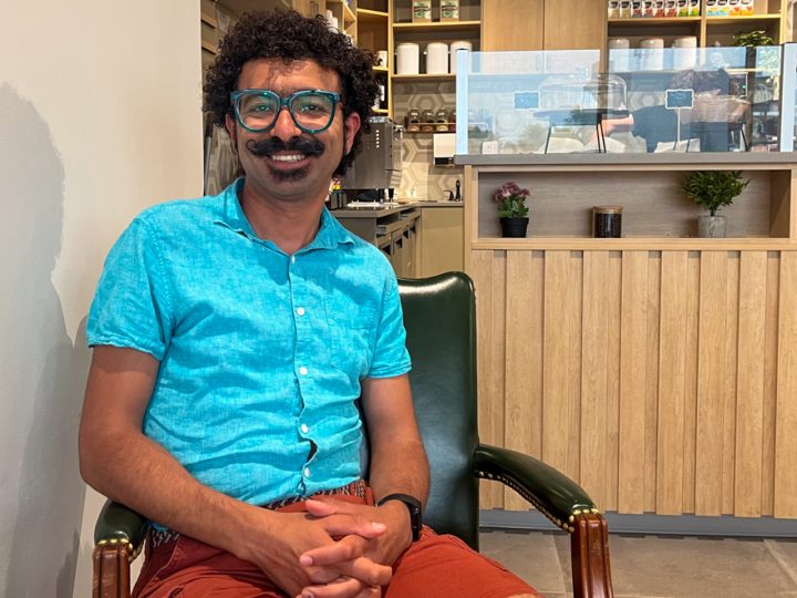 A brown-skinned man with glasses, black curly hair and a moustache is sitting on a chair in a coffee shop smiling. He is wearing a teal shirt and light red shorts.