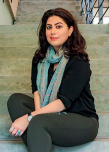 A woman sits at the bottom of a staircase wearing grey pants, a black top, and a turquoise scarf.