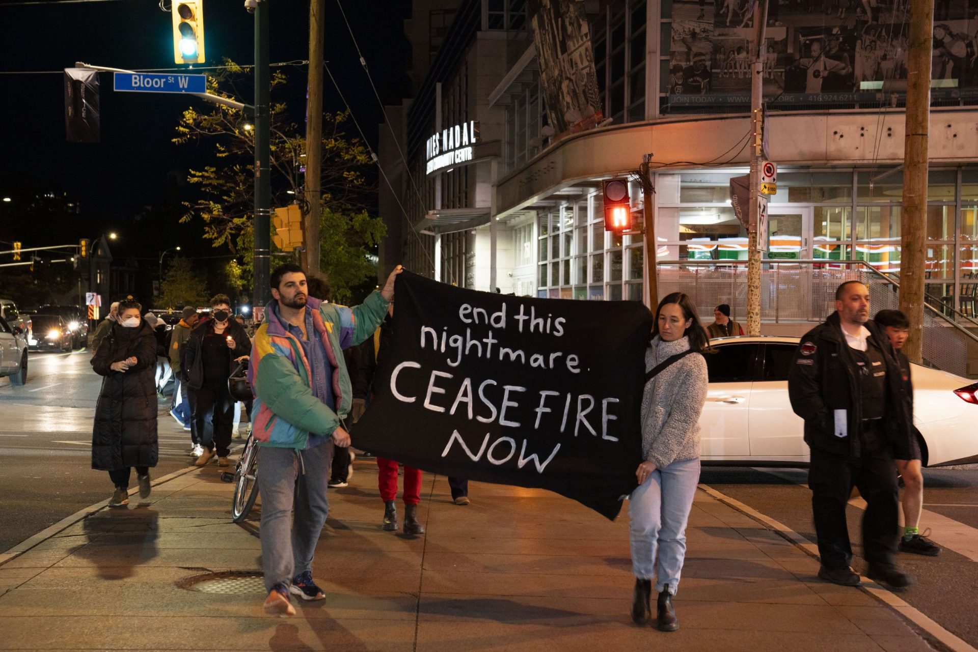 Two people in front of a crowd, walking along crosswalk with a black sign that says "End this nightmare. Ceasefire now."
