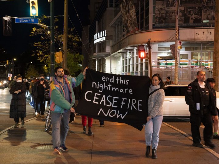 Two people in front of a crowd, walking along crosswalk with a black sign that says "End this nightmare. Ceasefire now."