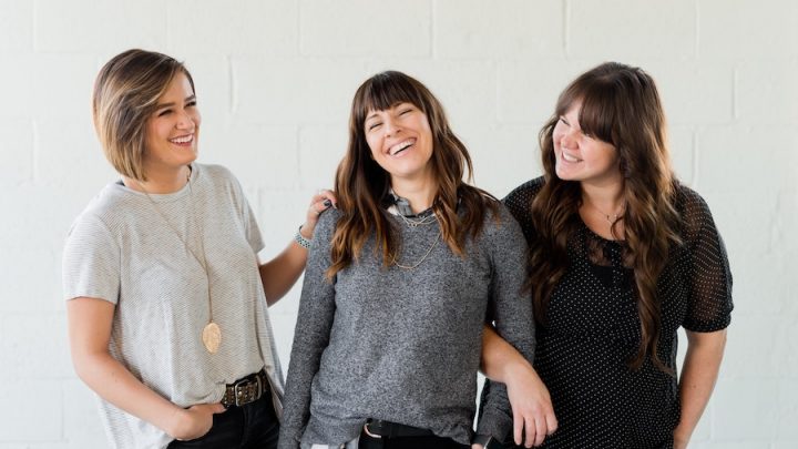 Three women smiling together. The first woman has short blond hair and is dressed in a light grey shirt and black pants. She is placing her hand on the second woman with shoulder-length brown hair, dressed in a dark grey shirt and black pants. Her arm is being held by the third woman with long brown hair, dressed in a black shirt and black pants.