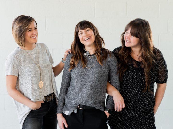 Three women smiling together. The first woman has short blond hair and is dressed in a light grey shirt and black pants. She is placing her hand on the second woman with shoulder-length brown hair, dressed in a dark grey shirt and black pants. Her arm is being held by the third woman with long brown hair, dressed in a black shirt and black pants.