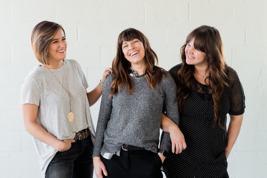 Three women smiling together. The first woman has short blond hair and is dressed in a light grey shirt and black pants. She is placing her hand on the second woman with shoulder-length brown hair, dressed in a dark grey shirt and black pants. Her arm is being held by the third woman with long brown hair, dressed in a black shirt and black pants.